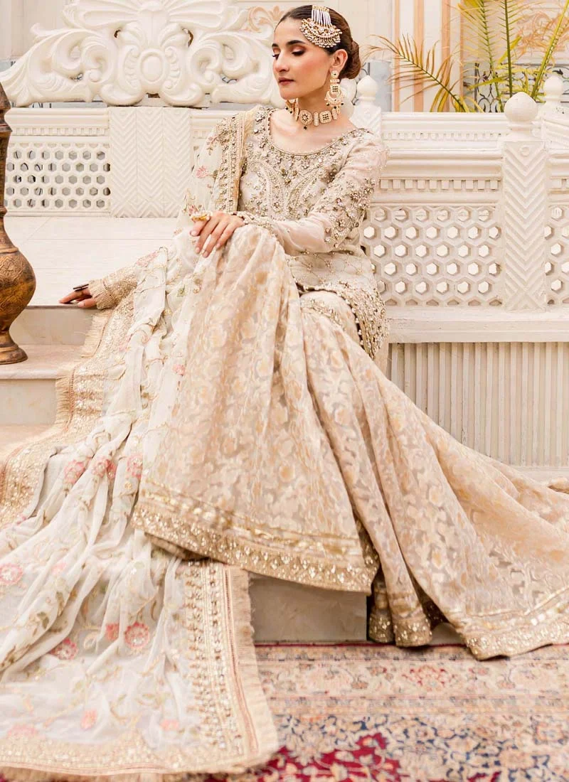 Ivory gharara suit seated view with wide patterned flares and gold-bordered floral dupatta